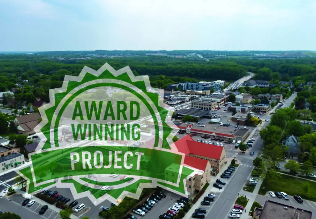 Aerial view of a small town with trees, roads, and buildings. Overlaid is a green and white badge with the words AWARD WINNING PROJECT in bold capital letters.