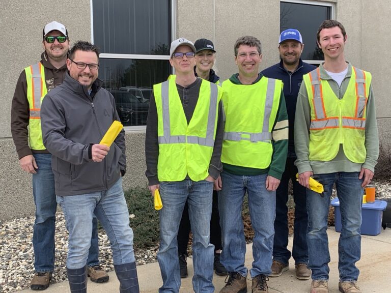 Seven people stand outdoors in front of a building, some wearing neon safety vests and holding yellow objects. Smiling in casual clothing, hats, and sunglasses.