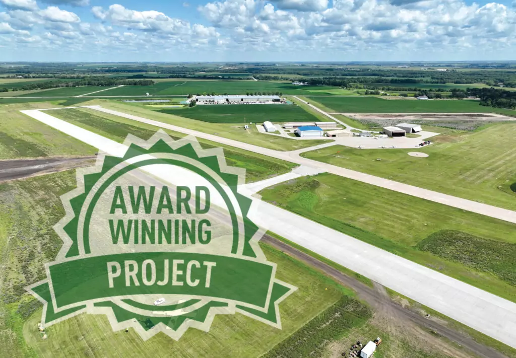 Aerial view of a rural airport with green fields and runways. A transparent badge graphic labeled “Award Winning Project” is overlaid on the image.