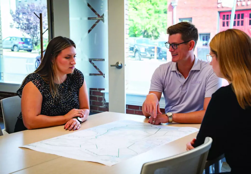 Three people sit around a table discussing a large map or blueprint. They are in a bright, modern office with glass walls and large windows.
