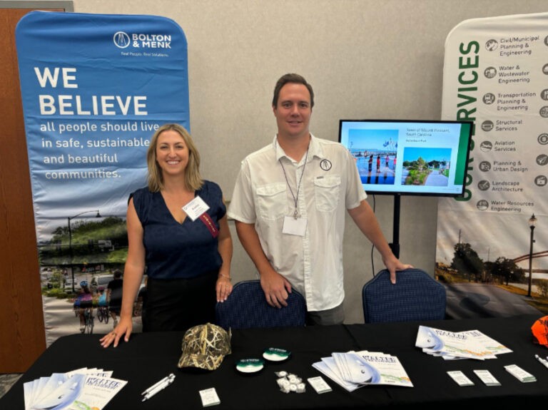 Two people stand behind a booth with brochures, business cards, and branded items. A display reads We Believe all people should have Real Opportunity to live in safe, sustainable, and beautiful communities. A TV and informational banners are in the background.
