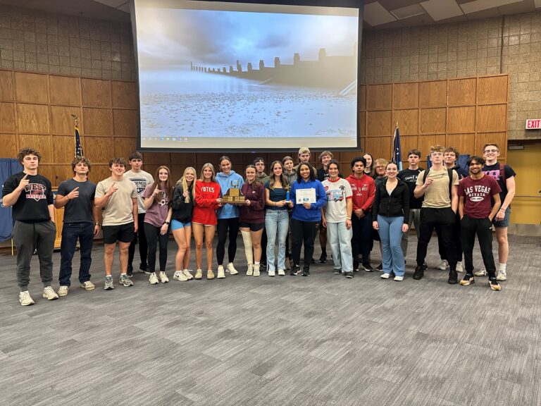 A group of young adults stands together indoors in front of a large projector screen, smiling at the camera. Some hold plaques or certificates celebrating their achievements in STEAM Education. The room has a gray carpet and tan walls.