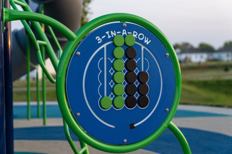 A close-up of a blue 3-in-a-row game board with green and black sliding discs arranged vertically and horizontally. Playground equipment and grass are visible in the background.