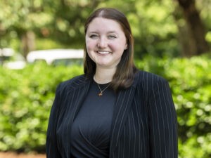 A woman with straight brown hair, wearing a black pinstripe blazer and black top, stands outdoors in front of green foliage, smiling at the camera.