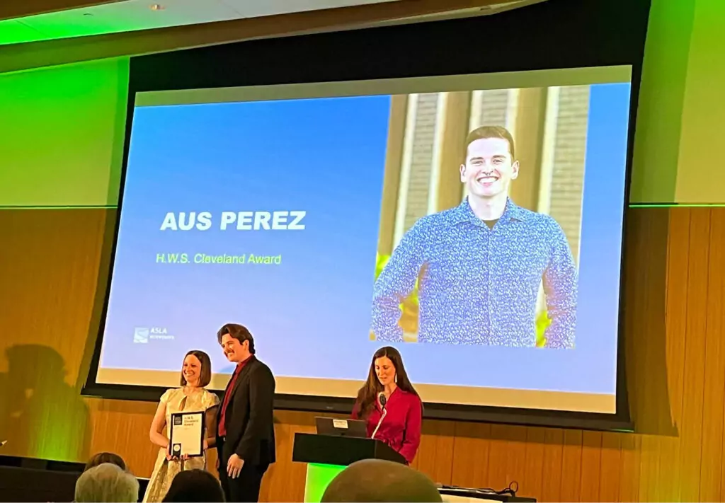 A man receives the Award on stage with two women. Behind them, a large screen shows a photo of a smiling man and the text Aus Perez, H.W.S. Cleveland Award.