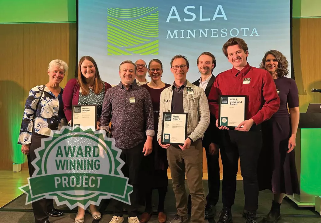 Eight people stand on a stage smiling, holding award plaques, with a screen behind them displaying the ASLA Minnesota logo. A green badge in the corner reads Award Winning Project.