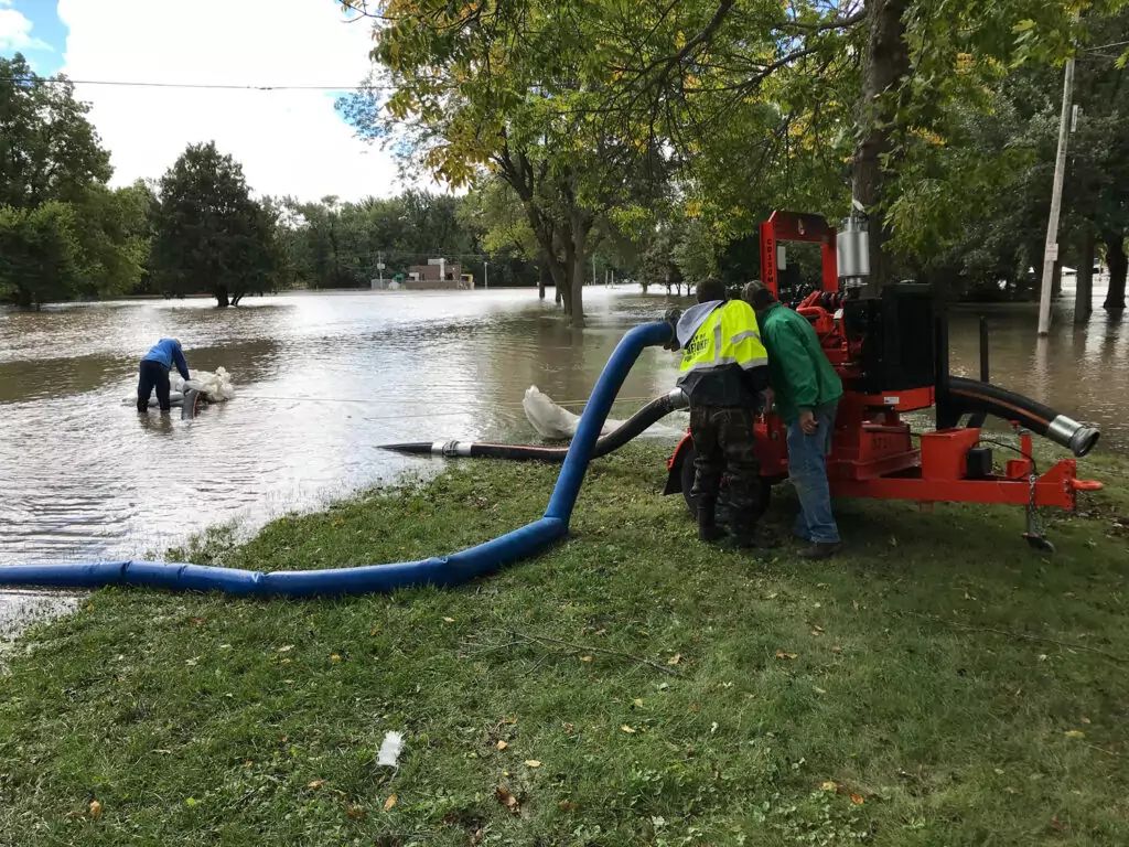 The image depicts a scene of flooding in a grassy area with several trees. Two individuals are working with a large red pump machine connected to blue hoses.
