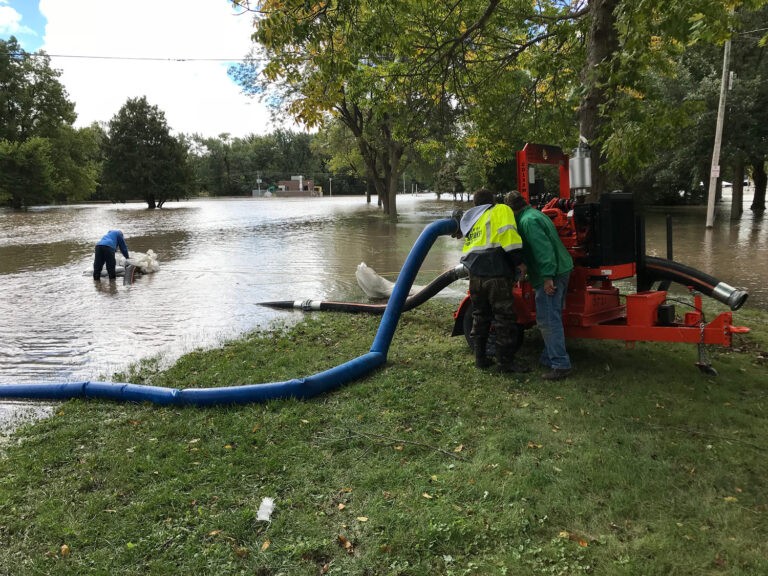 The image depicts a scene of flooding in a grassy area with several trees. Two individuals are working with a large red pump machine connected to blue hoses.