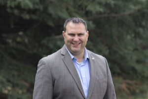 The image shows a man wearing a gray suit jacket and a light blue dress shirt standing outdoors in front of greenery.