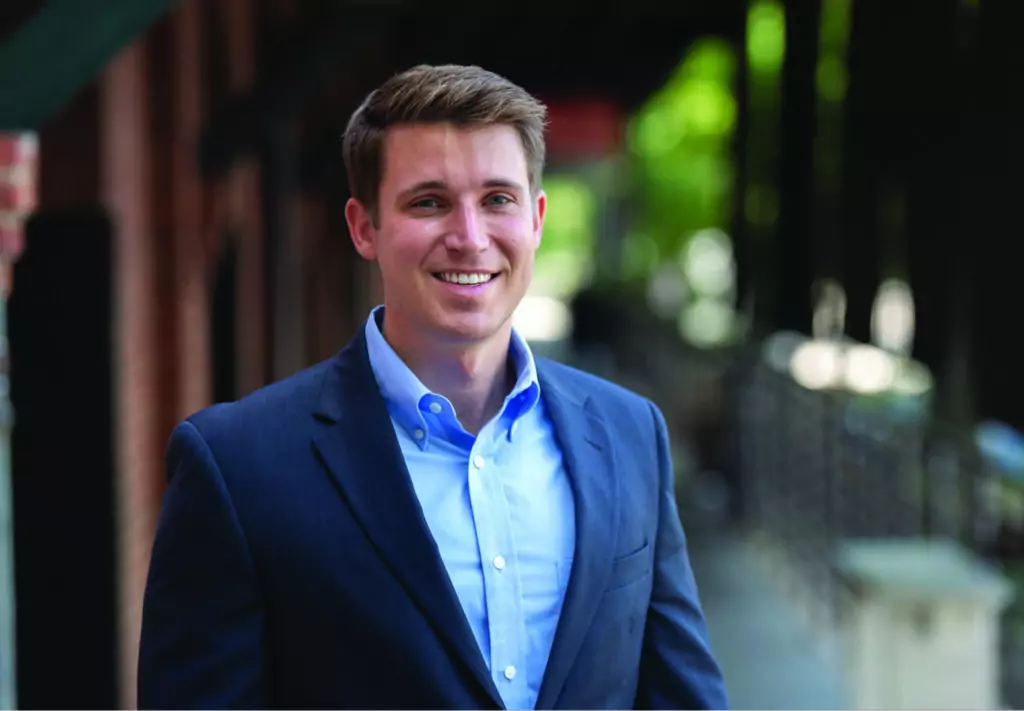 A man in a blue suit and light blue shirt smiles while standing outdoors near a brick building with trees and a blurred pathway in the background.