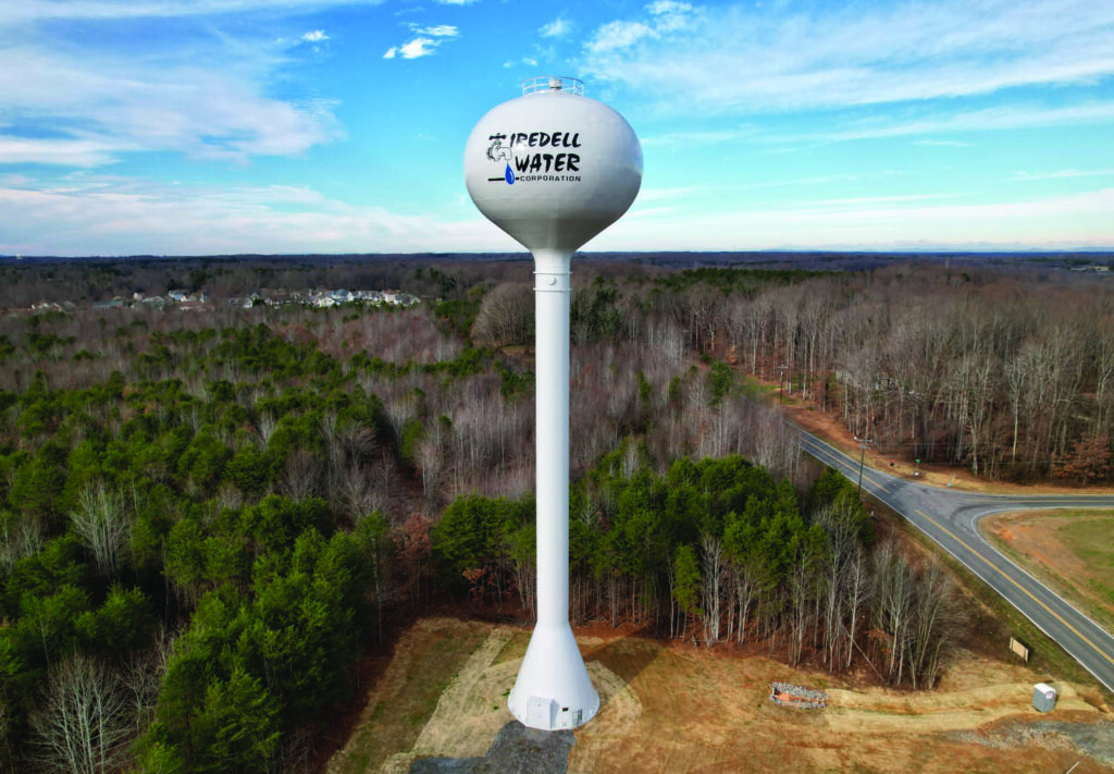 A tall white water tower labeled “Iredell Water Corporation” stands among trees near a curved road, with houses visible in the distance under a partly cloudy sky.