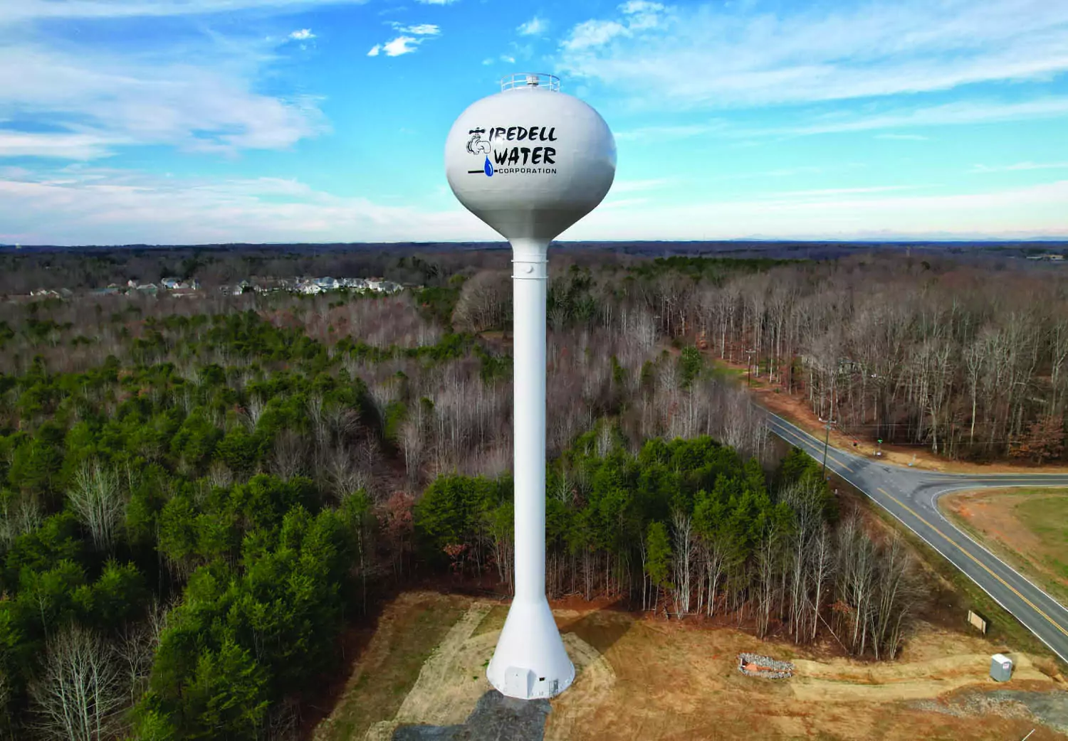 A tall white water tower labeled “Iredell Water Corporation” stands among trees near a curved road, with houses visible in the distance under a partly cloudy sky.