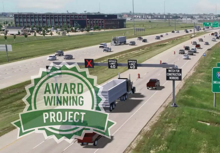 An aerial view of a busy highway with vehicles and digital speed limit signs, overlaid with a green Award Winning Project badge on the left side. Road signs and grassy areas are visible.