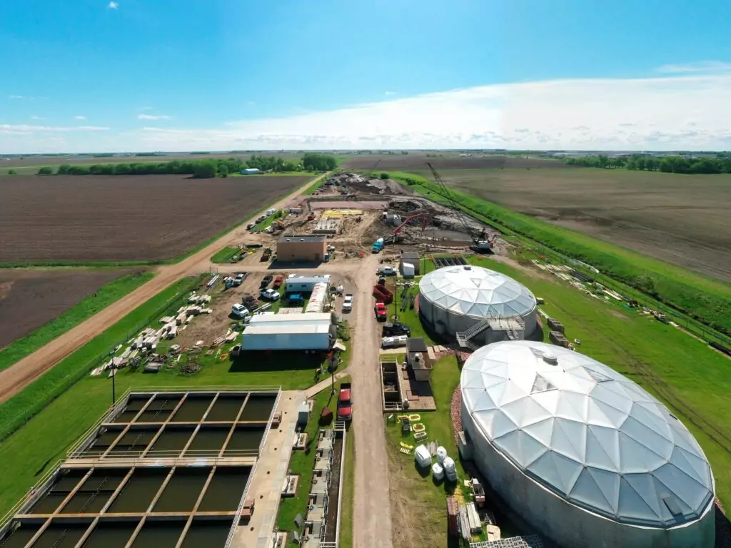 Aerial view of a rural construction site with large dome-shaped structures, buildings, equipment, and farmland stretching to the horizon under a blue sky.