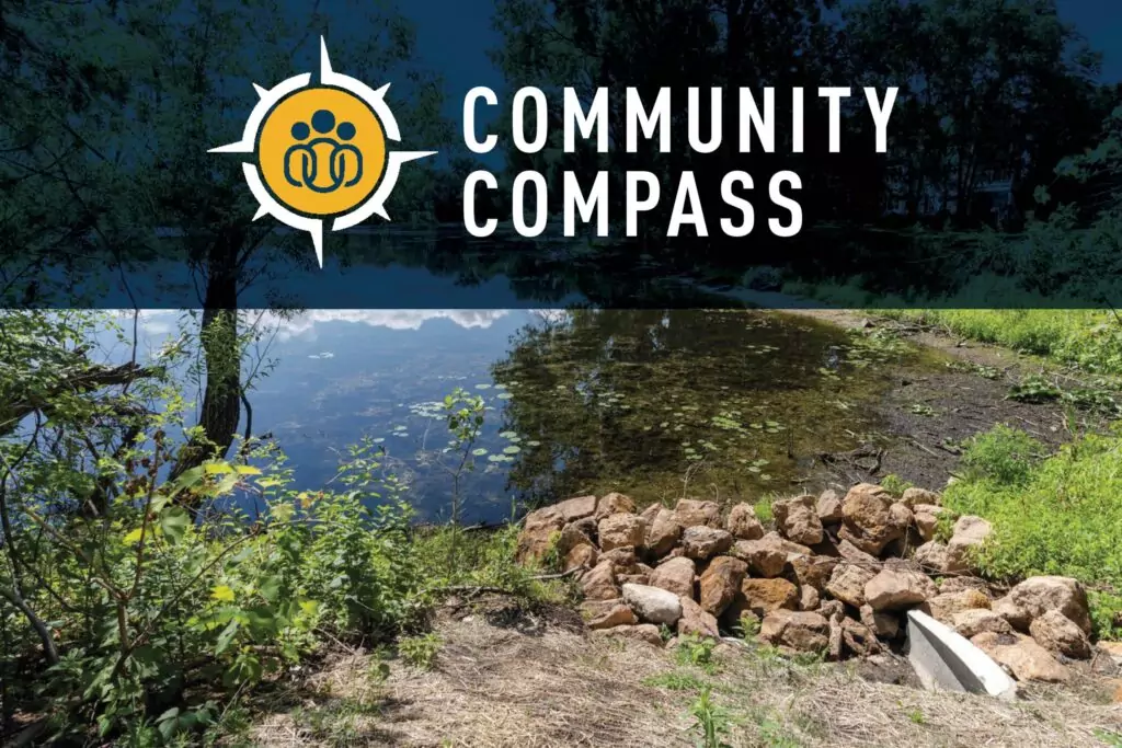A natural outdoor scene with a small pond surrounded by greenery. In the foreground, there is a pile of rocks and some vegetation. Trees and their reflections are visible in the background. At the top, a logo with a compass design and the text 'COMMUNITY COMPASS' is displayed