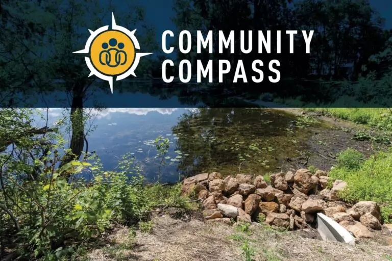 A natural outdoor scene with a small pond surrounded by greenery. In the foreground, there is a pile of rocks and some vegetation. Trees and their reflections are visible in the background. At the top, a logo with a compass design and the text 'COMMUNITY COMPASS' is displayed