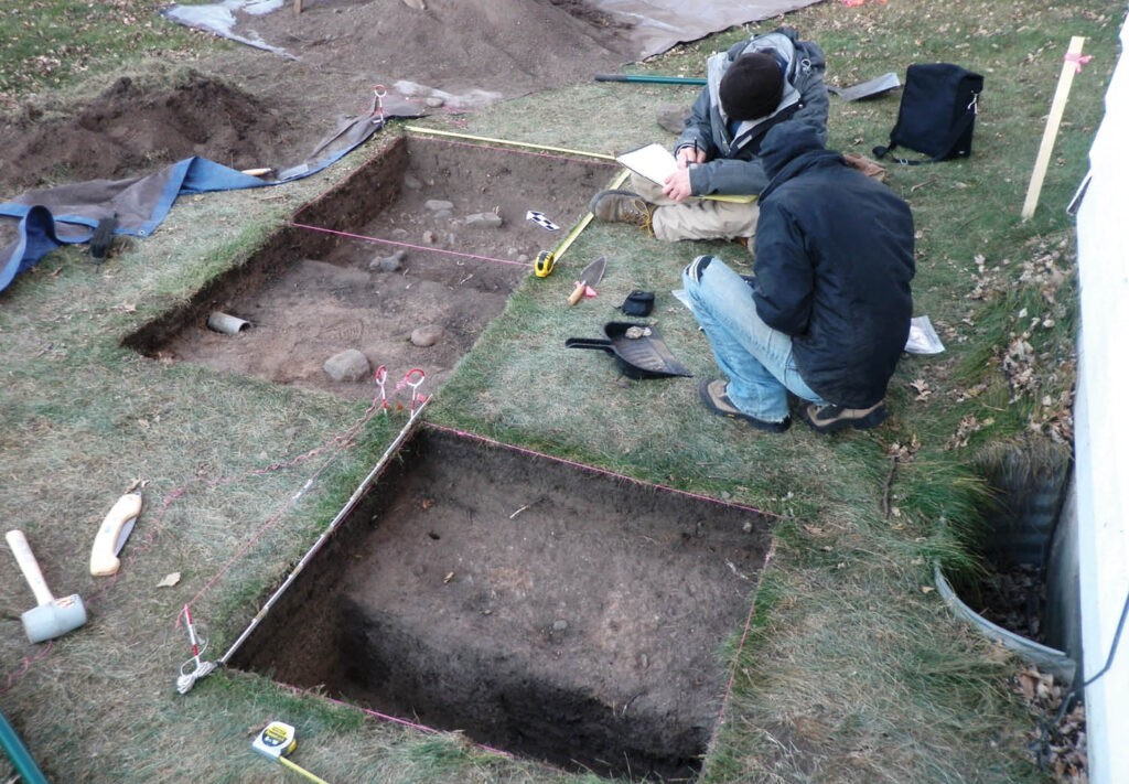Two people wearing jackets kneel beside rectangular excavated pits in grassy ground, taking notes and examining tools. The area is marked with string, and excavation tools are scattered nearby.