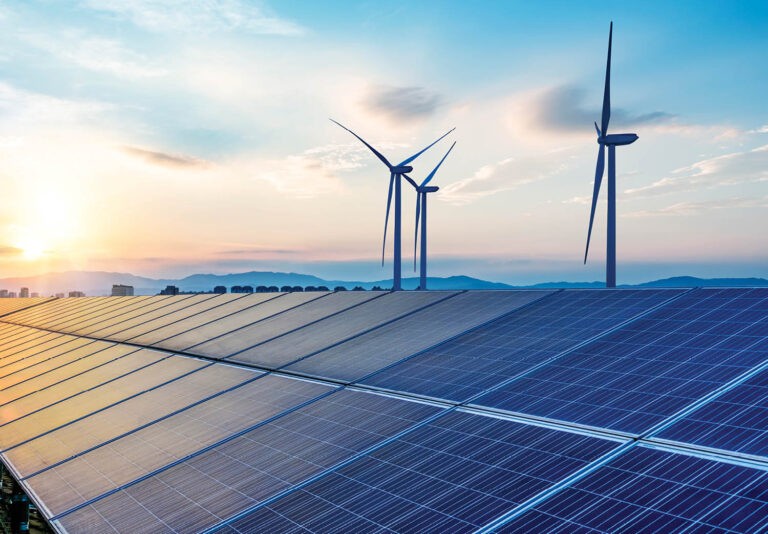 Solar panels and wind turbines generate renewable energy under a bright sky at sunset, with mountains visible in the background.