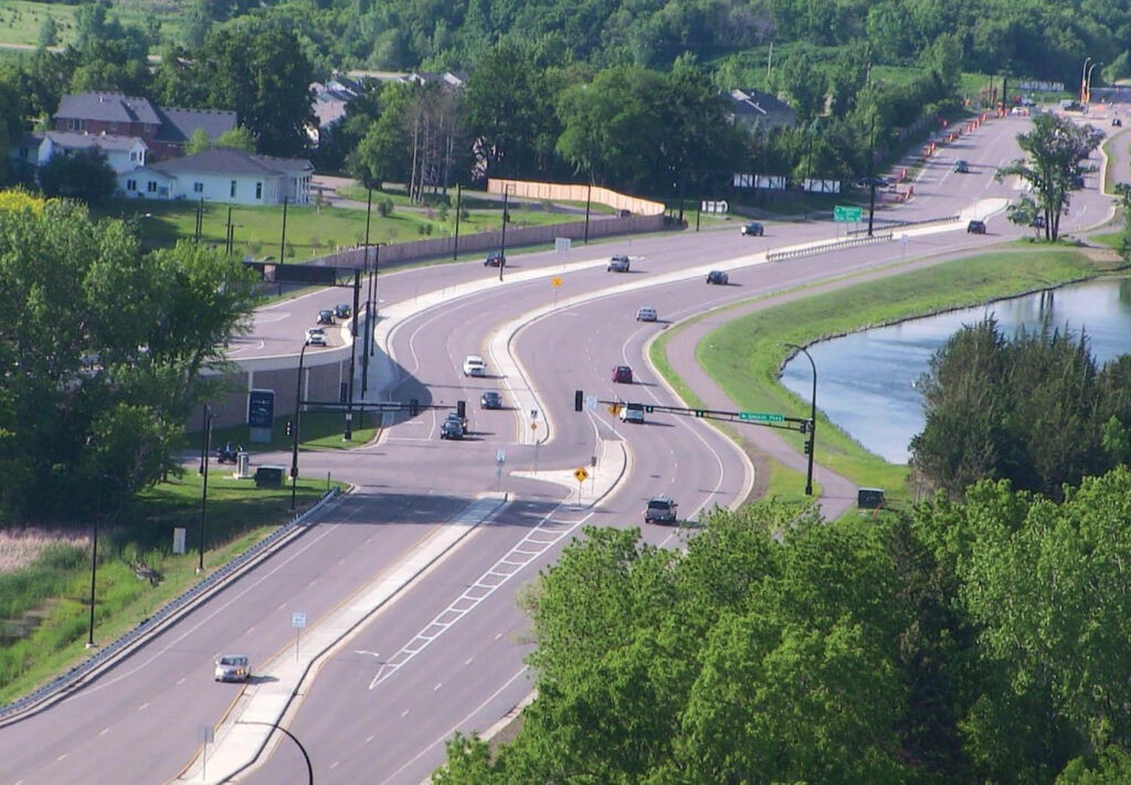 Aerial view of a multi-lane road with cars traveling in both directions, surrounded by greenery and trees, a body of water on the right, and houses in the background.