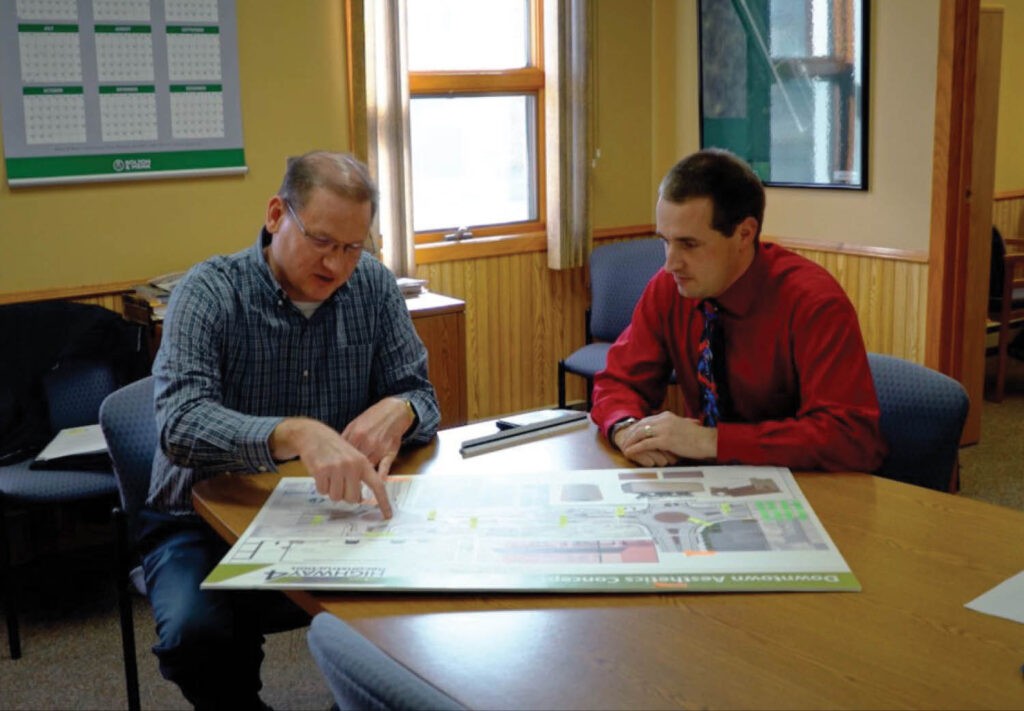 Two men sit at a table in an office, discussing and pointing at a large map or blueprint spread out in front of them. The room has wood paneling, windows, and a wall calendar.