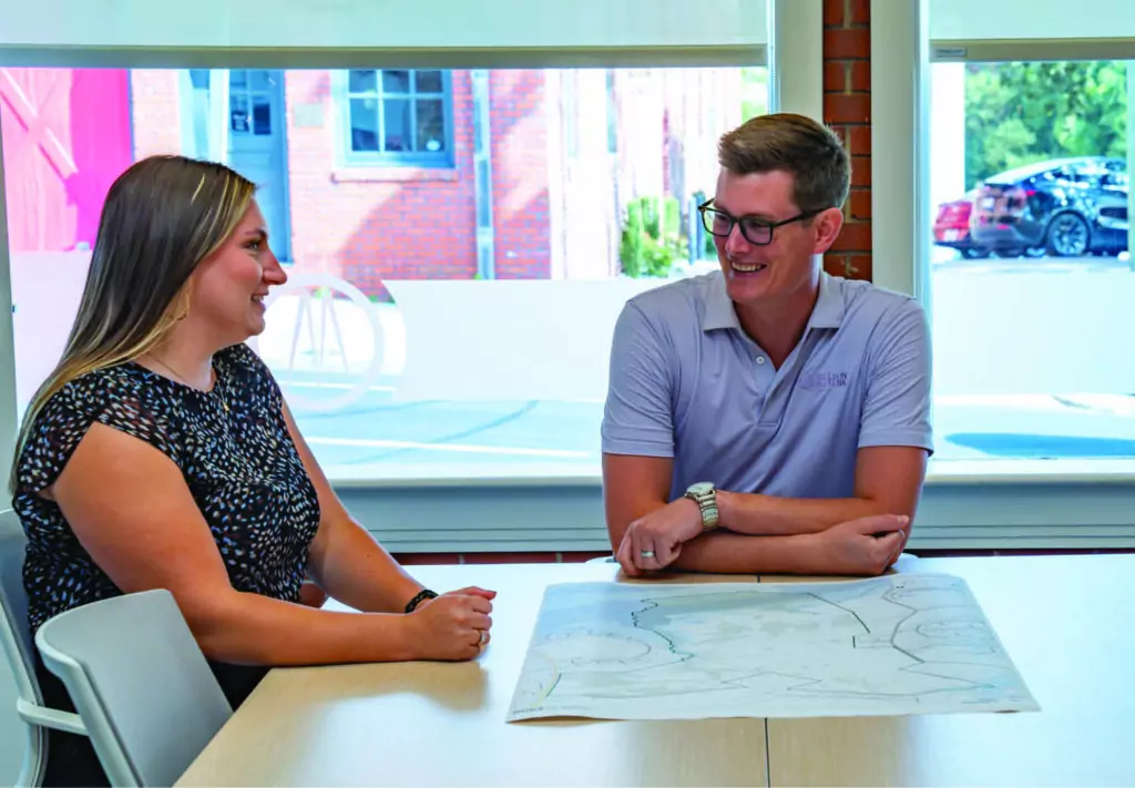 Two people sit at a table in a bright office, smiling and talking. A large paper with plans or drawings is spread out in front of them. Large windows reveal a colorful building and a parked car outside.
