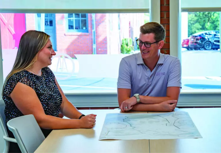 Two people sit at a table in a bright office, smiling and talking. A large paper with plans or drawings is spread out in front of them. Large windows reveal a colorful building and a parked car outside.