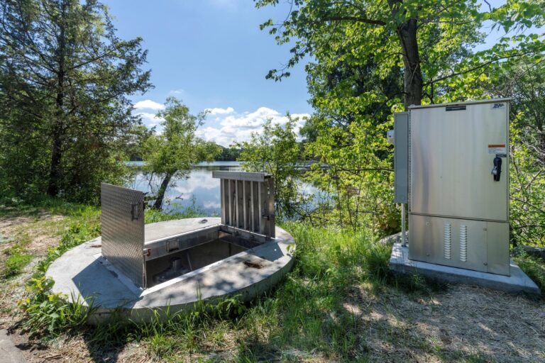 An outdoor scene featuring a body of water in the background, surrounded by trees and greenery. In the foreground, there is an open metal hatch on a concrete base to the left and a closed metal cabinet with visible controls or equipment to the right.