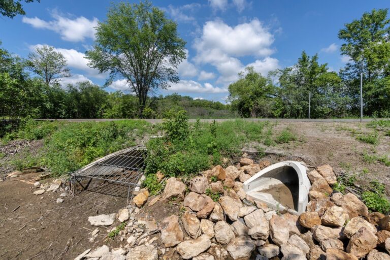 A culvert with two openings surrounded by rocks and vegetation; one opening is covered with a metal grate, and the other is an open concrete pipe. A road, trees, and a blue sky with scattered clouds are visible in the background.