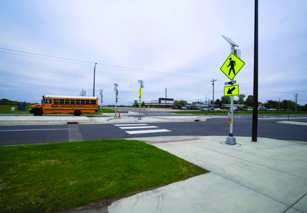 A crosswalk with pedestrian crossing signs and solar panels near a school bus stopped at the curb; the scene is by a street with sidewalks, grass, and a parking lot in the background under a cloudy sky.
