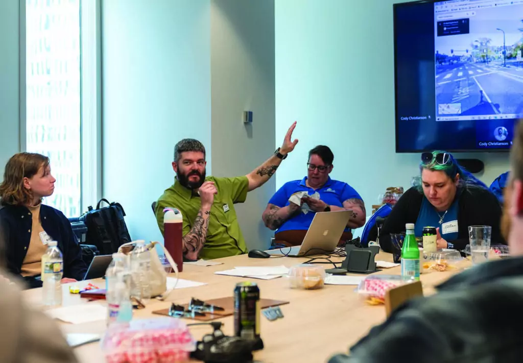 A group of people sit around a conference table with laptops, drinks, and papers. One person with tattoos raises their hand while speaking. A screen on the wall displays a street view map.