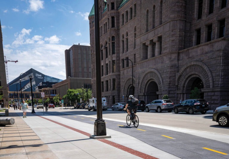 A person rides a bicycle in a bike lane beside a busy street lined with historic stone buildings, with a modern stadium visible in the background under a partly cloudy sky.