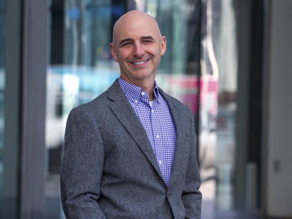 A smiling bald man wearing a grey blazer over a blue and white checkered shirt stands outside in front of a modern glass building.