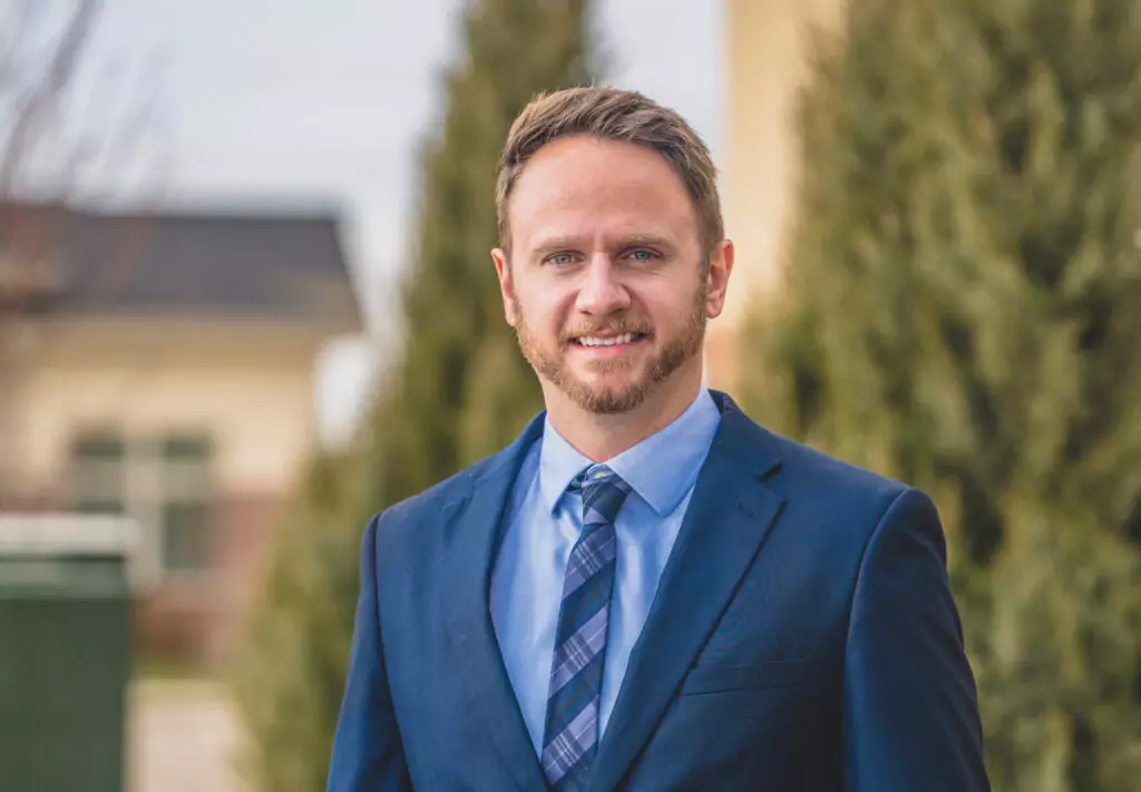 A man in a blue suit and tie stands outdoors, smiling at the camera. There are tall evergreen trees and blurred buildings in the background on a bright day.
