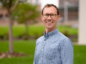 A man with short brown hair and glasses smiles while standing outdoors, perhaps after working on Reestablishing the River. He wears a light blue patterned button-up shirt, with a green, blurred park or garden background behind him.