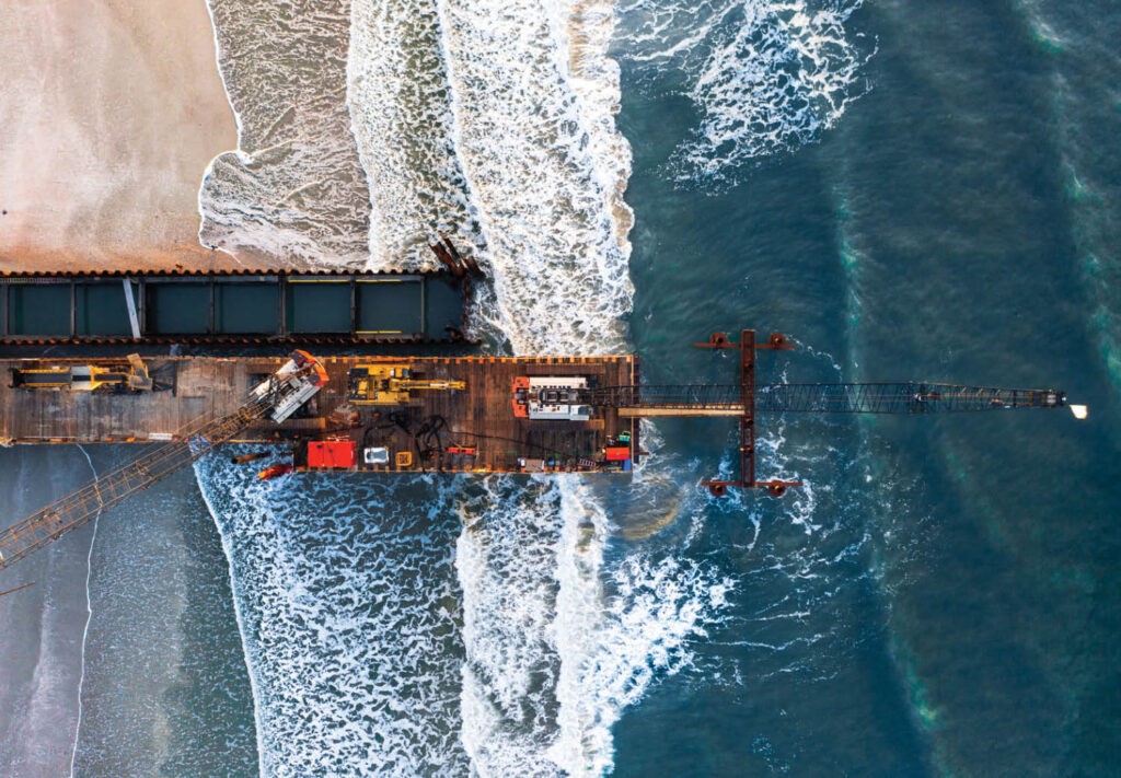 Aerial view of a construction platform extending from a sandy beach into the ocean, with waves crashing against the structure and equipment visible on the platform.