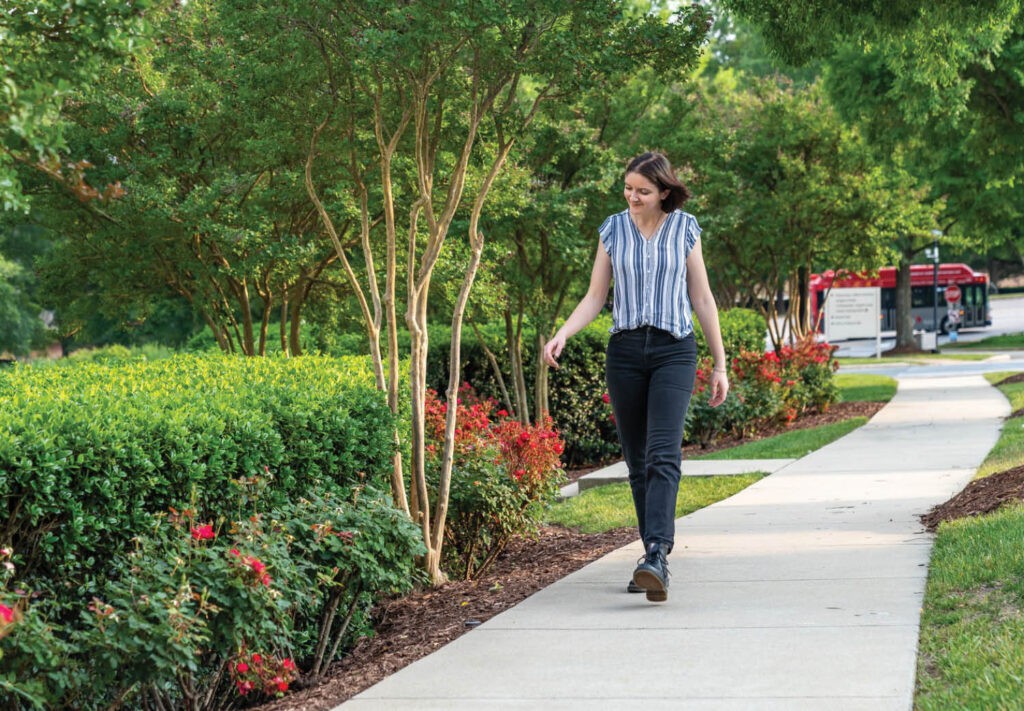 A woman in a striped shirt and black pants walks along a winding sidewalk surrounded by green bushes, red flowers, and trees on a sunny day.