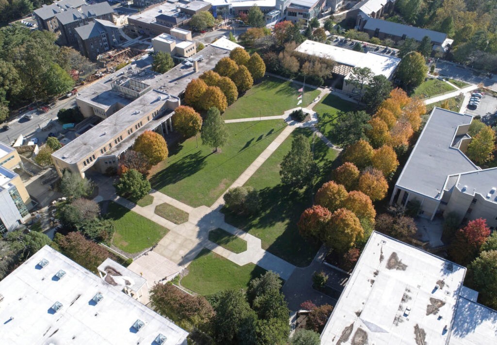 Aerial view of a green campus quad with diagonal walkways, surrounded by buildings and trees with autumn foliage, and a central circular area with a monument or statue.