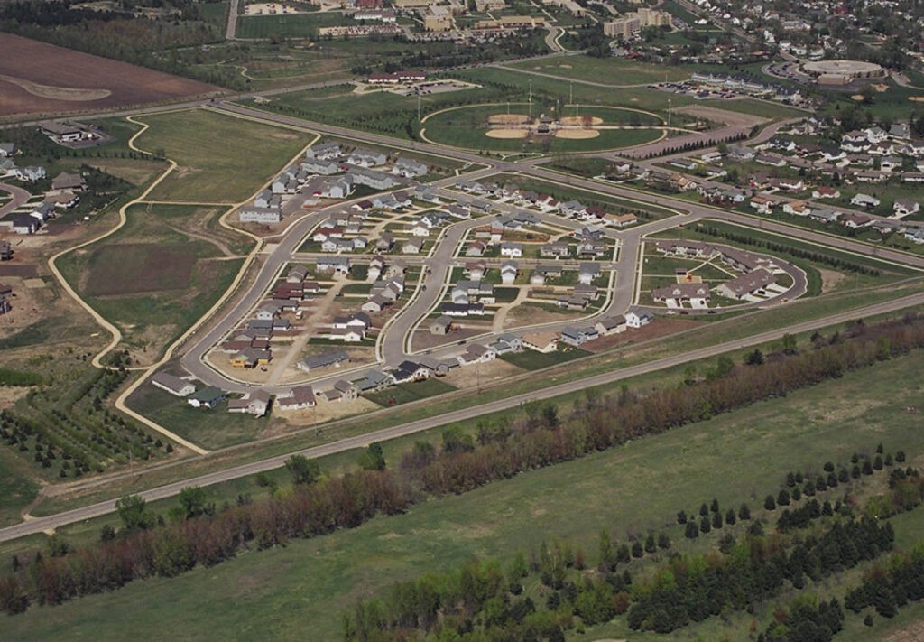 Aerial view of a suburban neighborhood under development, with winding roads, houses, and open plots of land. Surrounding areas include trees, fields, and a sports complex with baseball fields in the background.