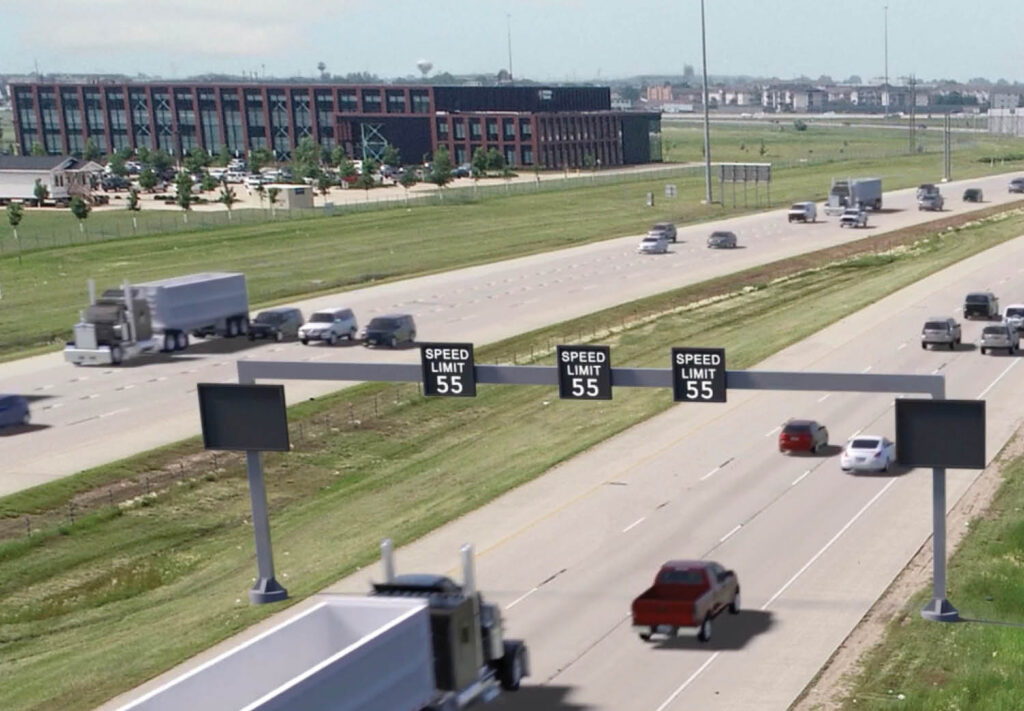 Cars and trucks travel on a multi-lane highway with electronic signs displaying a speed limit of 55 mph. Buildings, grass, and trees are visible in the background under a clear sky.