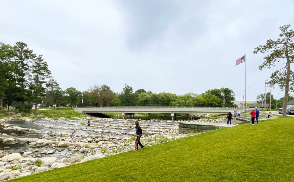 People walk along a grassy area beside a rocky stream, with a bridge, trees, and an American flag visible in the background under a cloudy sky.