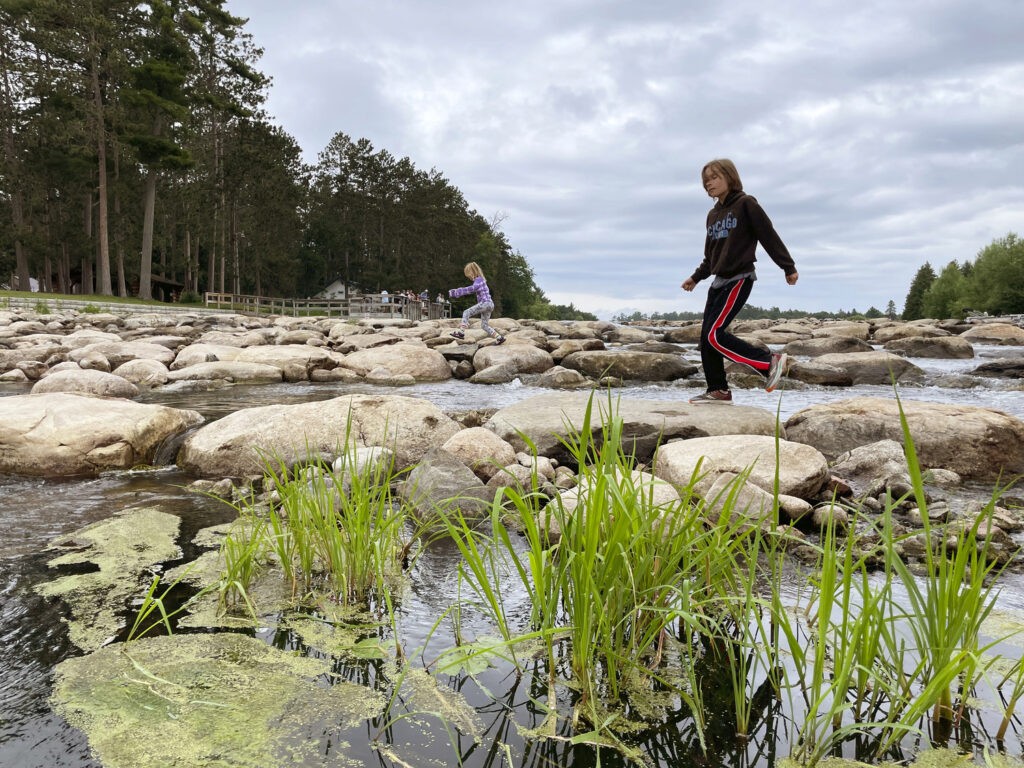Two girls are walking on large rocks across a shallow river, with green grass and algae in the foreground and tall trees in the background under a cloudy sky.