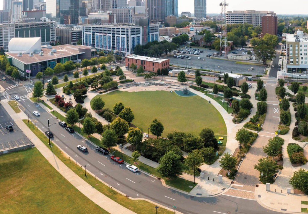 Aerial view of a circular city park with green lawns, trees, walking paths, and surrounding roads, set against an urban backdrop of buildings and parking lots on a sunny day.