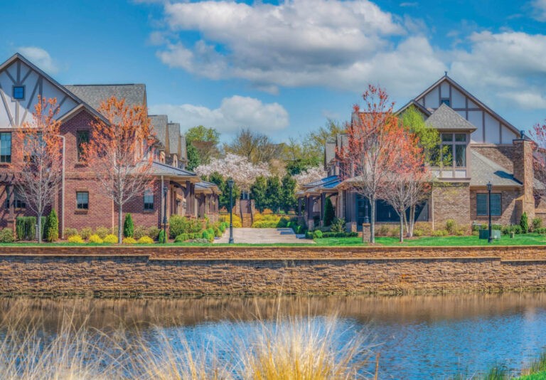A row of modern suburban houses with brick facades sits beside a calm pond, bordered by young trees with reddish leaves and landscaped greenery under a blue sky with scattered clouds.