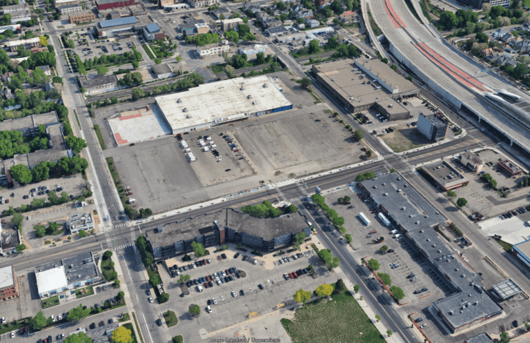 Aerial view of a city block highlighting large parking lots, several commercial buildings, and nearby roads—including a highway with overpasses—set against trees and residential areas poised for community revival in the background.