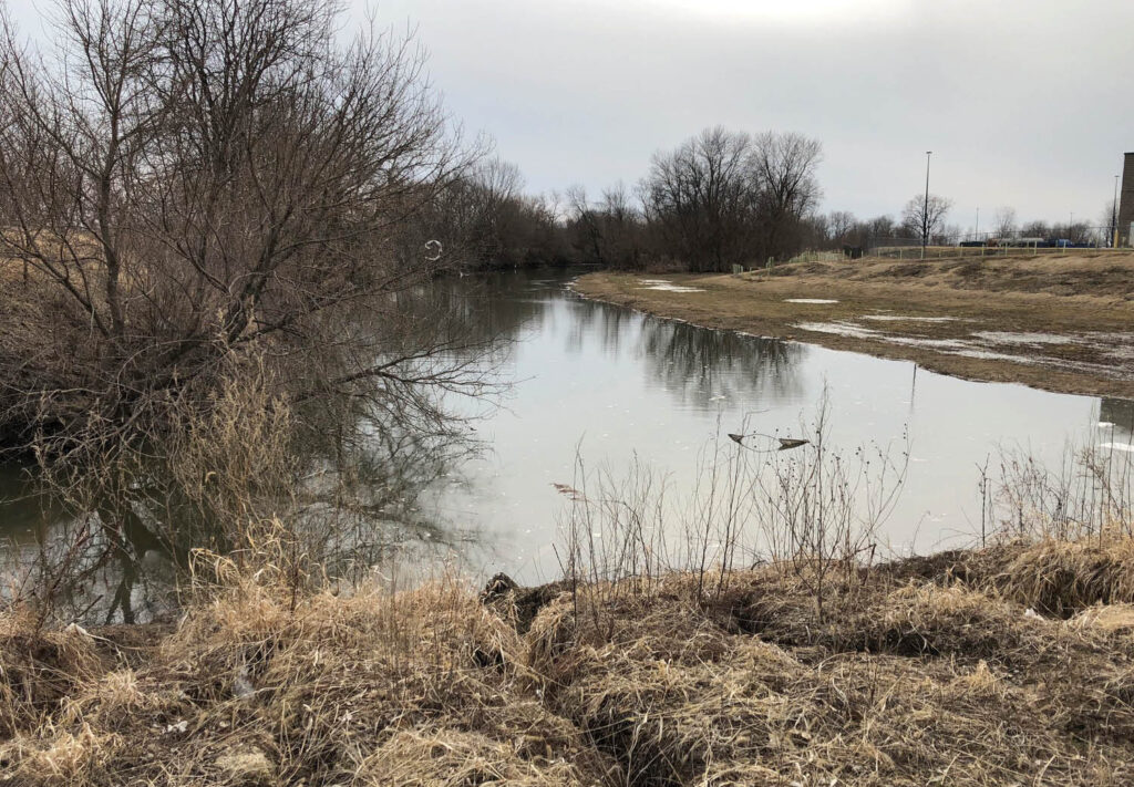 A calm, narrow river curves through a landscape of dry, brown grass and leafless trees under a cloudy sky in early spring or late winter. The water reflects the overcast sky and bare branches.