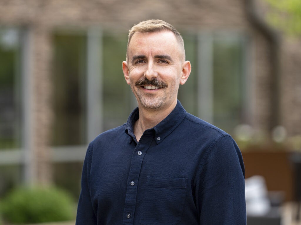 A man with short blond hair, a mustache, and a trimmed beard is smiling outdoors. He is wearing a navy blue button-up shirt. The blurred background of greenery and a brick building hints at vibrant community revival.