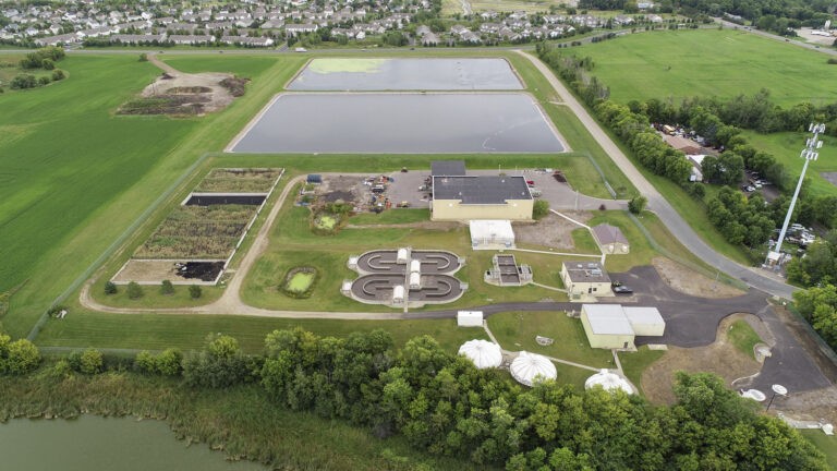 Aerial view of a wastewater treatment plant featuring sedimentation tanks, aeration tanks, clarifiers, and large rectangular treatment ponds, surrounded by green fields and a residential area in the background.