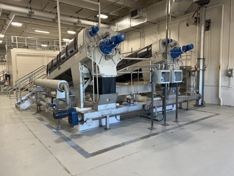 Industrial room with two large cylindrical machines featuring blue end caps, mounted on a metal framework, surrounded by pipes, valves, and control panels, with a high ceiling and exposed ductwork.