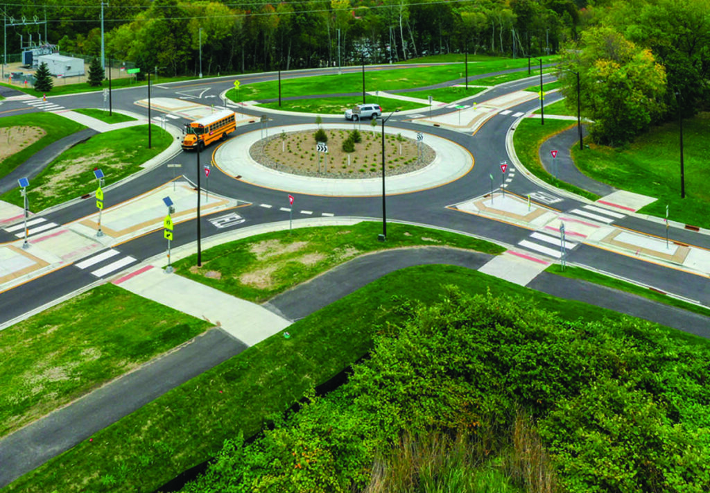 Aerial view of a large roundabout with landscaped center, surrounded by crosswalks, road signs, and greenery. A school bus, car, and pickup truck are navigating the intersection. Trees and grass border the area.