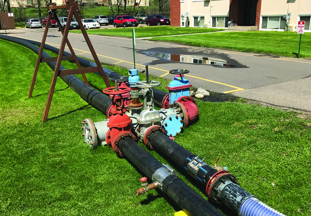 Pipes with multiple valves and connectors are arranged on a grassy area near a road, with cars parked in the background and a building visible across the street.